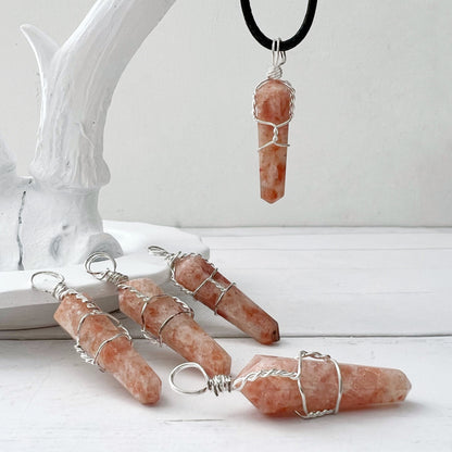 A 1.5" Double Terminated Sunstone Point Pendant consisting of an orange and white crystal wrapped in silver wire hangs from a black cord, evoking self-empowerment jewelry. The background is a plain white surface.