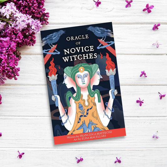 There is a book titled "Oracle of Novice Witches" lying on a white wooden surface, surrounded by sprigs of purple and white flowers. The cover features an illustration of a three-faced figure holding torches, with intertwining branches and crows in the background—a perfect companion for your divination tools.