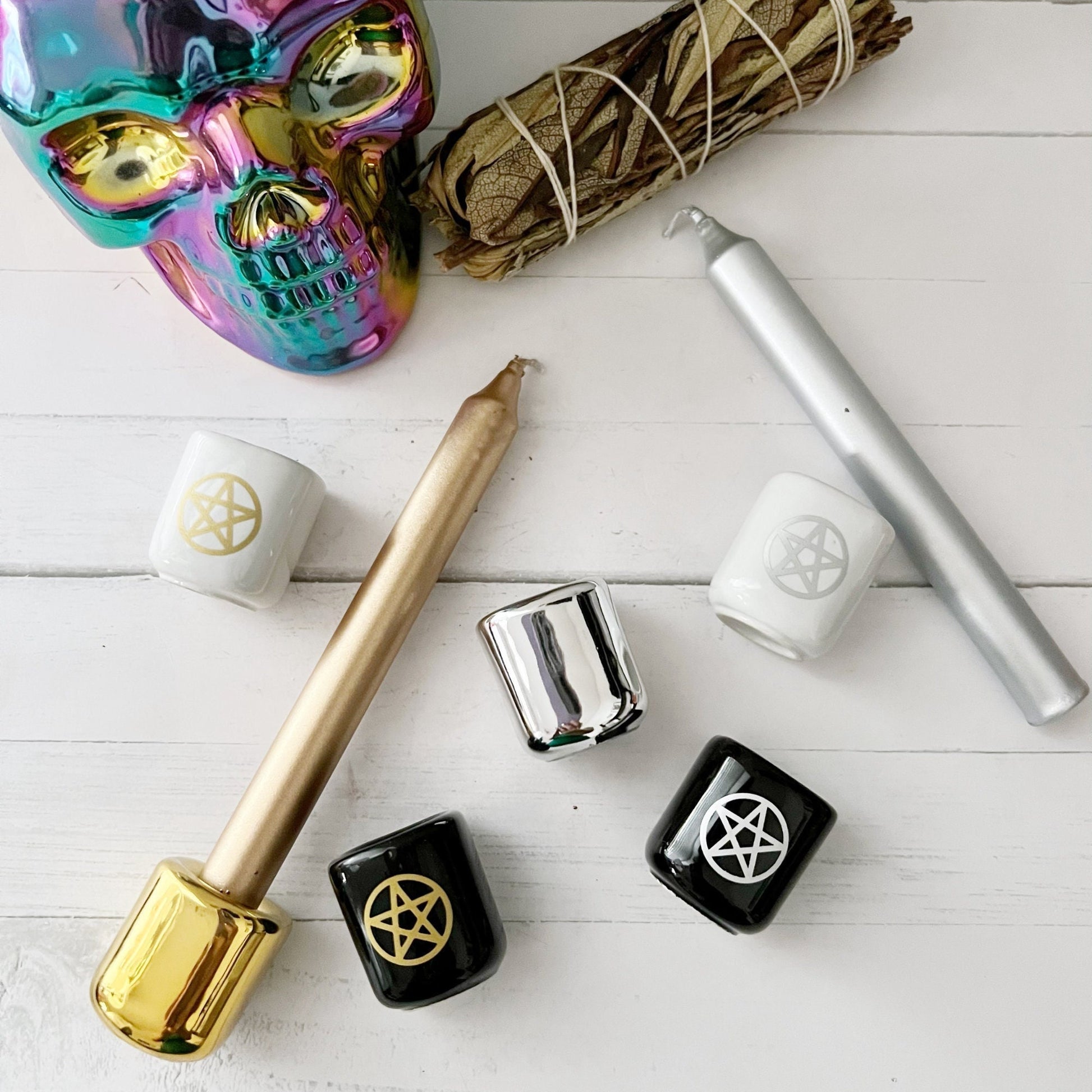 A collection of witchcraft tools on a white surface, including a multicolored metallic skull, sage bundle, gold and silver candles, several black, white, and silver candle holders decorated with pentagrams, and Pentacle Chime Ceramic Holders perfect for spellwork or altar decor.