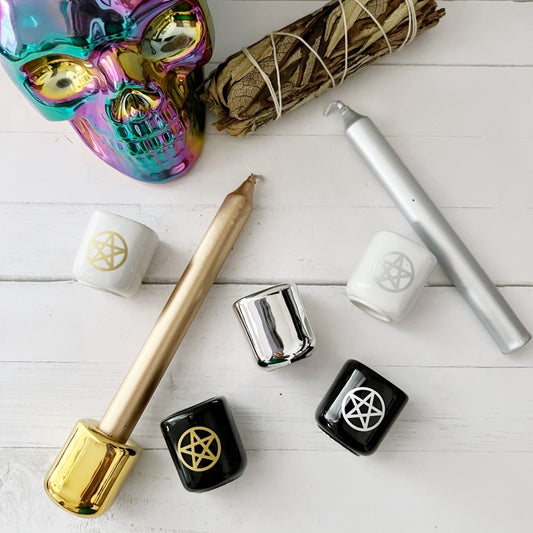 A collection of witchcraft tools on a white surface, including a multicolored metallic skull, sage bundle, gold and silver candles, several black, white, and silver candle holders decorated with pentagrams, and Pentacle Chime Ceramic Holders perfect for spellwork or altar decor.