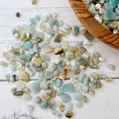 A wooden bowl filled with small, polished, multicolored stones, predominantly Amazonite Crystal Chips in blue and white, sits on a white wooden surface. Nearby, a bundle of tied sage, an energy filter in a clear plastic bag, and several small glass jars of stones are arranged for display.