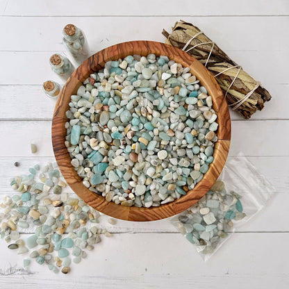 A wooden bowl filled with small, polished, multicolored stones, predominantly Amazonite Crystal Chips in blue and white, sits on a white wooden surface. Nearby, a bundle of tied sage, an energy filter in a clear plastic bag, and several small glass jars of stones are arranged for display.