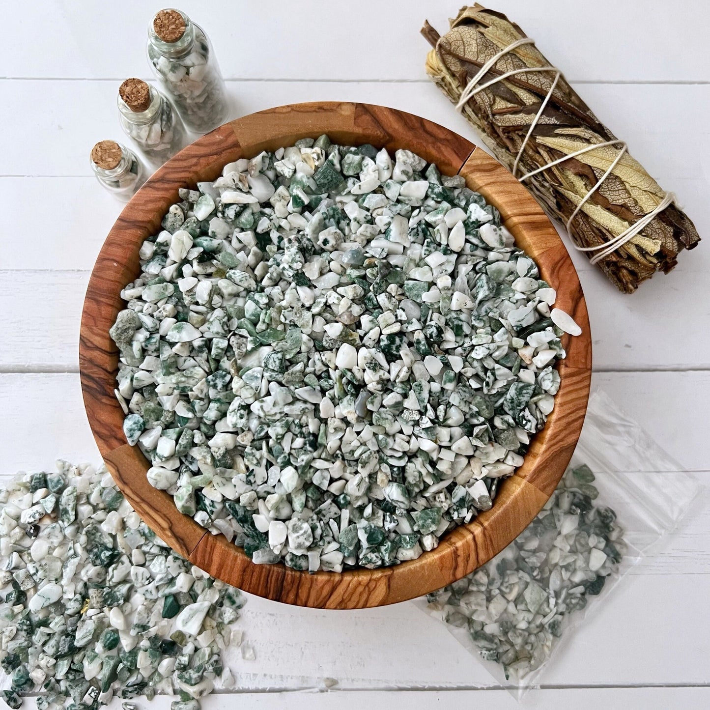 A wooden bowl filled with white and green Tree Agate Crystal Chips, set on a white surface. To the left of the bowl, small crystal chips are spilled on the table. On the upper left, two small corked bottles contain more crystal chips, and on the right, a sage bundle is wrapped with string.