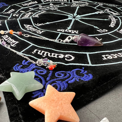 A black astrology board, akin to a 12" x 12" Embroided Velvet Zodiac Pendulum Mat, rests on the table, surrounded by small pumpkins and colorful potion bottles. This divination tool features a zodiac wheel with responses like "Yes," "No," and "Maybe," along with days of the week and calendar months.