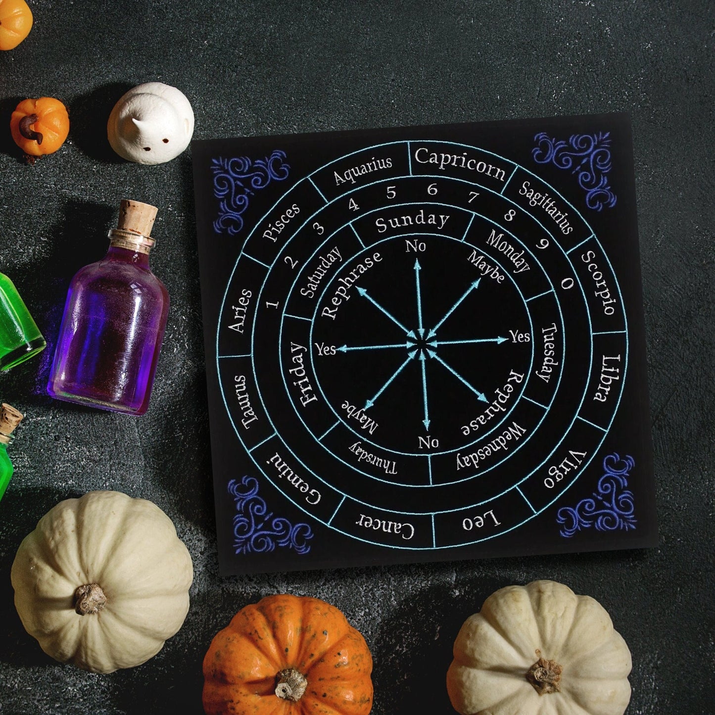 A black astrology board, akin to a 12" x 12" Embroided Velvet Zodiac Pendulum Mat, rests on the table, surrounded by small pumpkins and colorful potion bottles. This divination tool features a zodiac wheel with responses like "Yes," "No," and "Maybe," along with days of the week and calendar months.