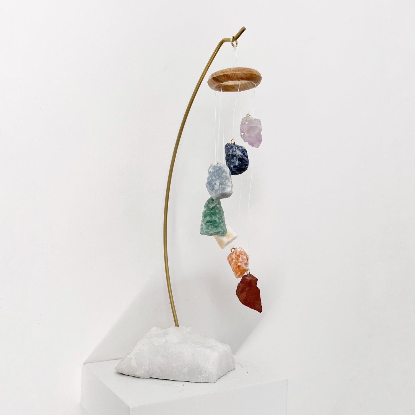 A decorative hanging display features a 7 Chakra Crystal Table Chime suspended on strings from a wooden ring, held by a curved metal stand anchored in a white stone base. Nearby are two potted succulent plants, one in a white pot and the other in a wooden container.