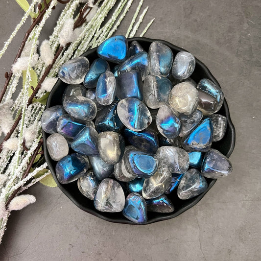 A black bowl is filled with polished, iridescent stones displaying a mix of blue, gray, and white hues. The bowl, featuring Indigo Aura Quartz Tumbled Stones believed to enhance psychic abilities, is placed on a gray surface beside a decorative arrangement of frosted twigs and leaves.