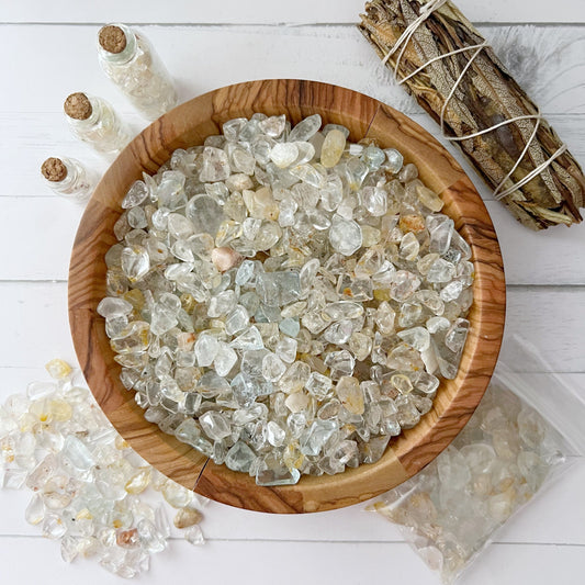 A wooden bowl filled with clear and slightly colored crystal stones sits on a white wooden surface. Nearby, small glass vials filled with crystals and an open bag of Natural Topaz Crystal Chips spill out, accompanied by a sage smudge stick wrapped in twine—a perfect setup for metaphysical crafting.