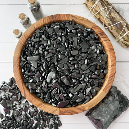 A wooden bowl filled with polished black stones sits on a white wooden surface. Surrounding the bowl are small glass bottles filled with stones, a bundle of sage tied with string, a plastic bag of Shungite Crystal Chips for EMF protection, and scattered stones on the surface.