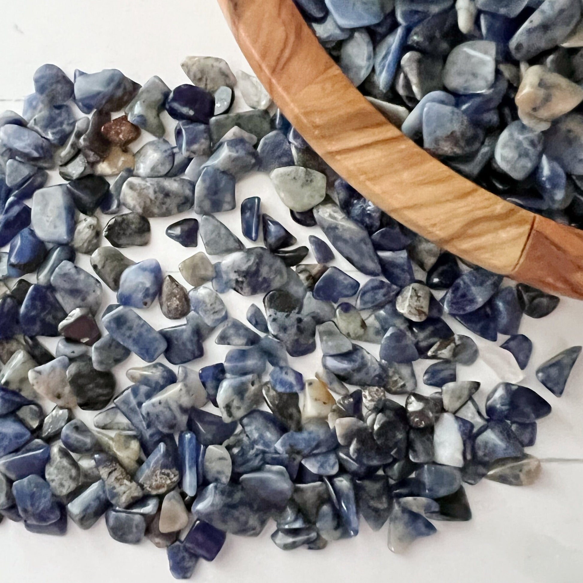 A wooden bowl filled with small blue Sodalite Crystal Chips sits on a white surface. Small jars containing stones are placed nearby, along with a bundle of sage for smudging. Additional stones are scattered around the bowl and a bag of Sodalite Crystal Chips is also visible, enhancing psychic abilities during meditation.