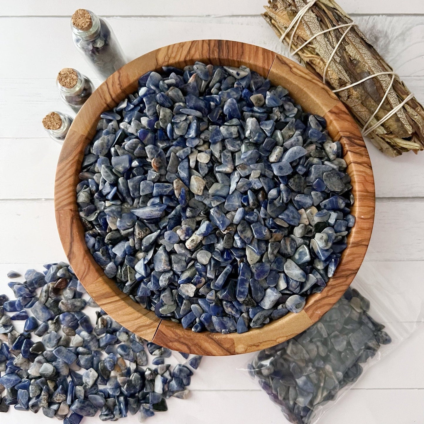 A wooden bowl filled with small blue Sodalite Crystal Chips sits on a white surface. Small jars containing stones are placed nearby, along with a bundle of sage for smudging. Additional stones are scattered around the bowl and a bag of Sodalite Crystal Chips is also visible, enhancing psychic abilities during meditation.