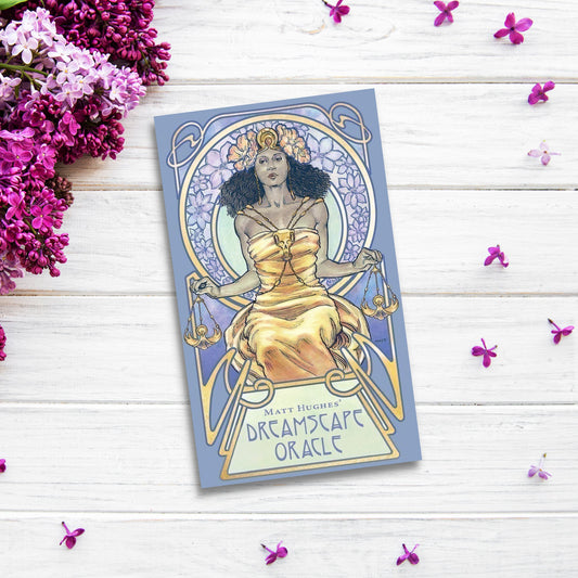 Dreamscape Oracle placed on a light wooden surface, surrounded by sprigs of purple flowers. This divination deck features an illustration of a woman in a golden dress holding scales, with a decorative, ornate border.