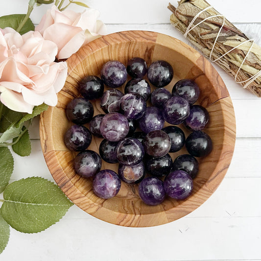 A wooden bowl filled with polished black and purple stones, including Mini Dream Chevron Amethyst Spheres, is placed on a white surface. To the left of the bowl, there are pink roses and green leaves. In the top right corner, a bundle of sage wrapped in string awaits your meditation or energy healing sessions.