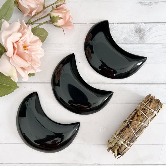 The image shows three 4" Obsidian Crescent Moons arranged in a line on a white wooden surface. To the left, there are soft pink flowers with green leaves. To the right, a smudge stick wrapped in white thread is placed on the surface.