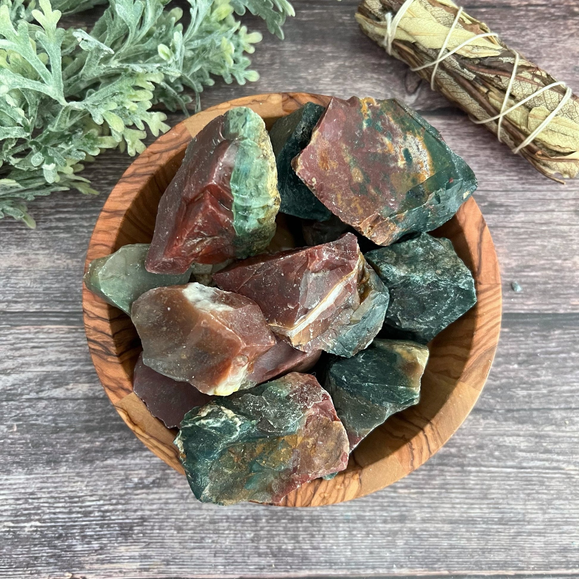 A wooden bowl filled with rough, multicolored Raw Fancy Jasper Stones sits on a wooden surface. Behind the bowl are green leaves and a bundle of sage tied with string. The calming stones display a variety of red, green, and brown hues with varied textures, embodying the Stone of Tranquility.