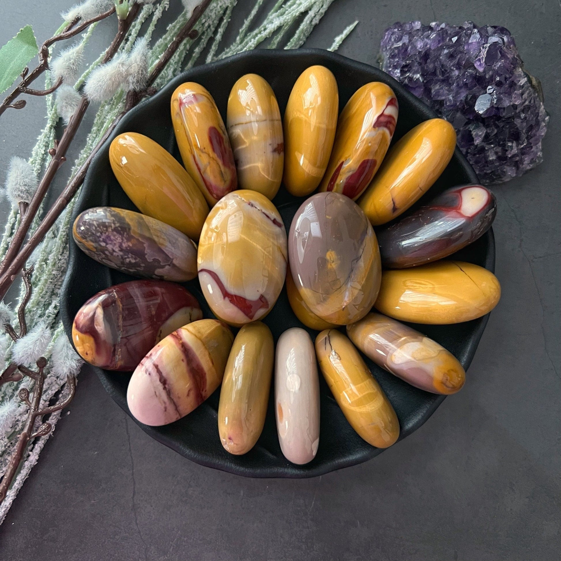 A black dish holds an array of polished, oval-shaped stones of various colors, including shades of brown, yellow, and red. Among them is a Mookaite Palm Stone radiating rejuvenating earth energy. The dish is placed on a dark surface with frosted branches and a purple crystal cluster nearby.