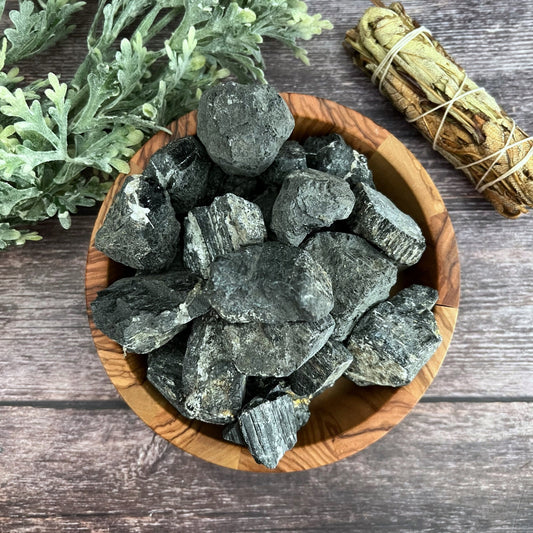 A wooden bowl filled with rough, dark stones, including Raw Black Tourmaline Stones known for their grounding properties, sits on a wooden surface. Next to the bowl, there is a bundle of sage wrapped in twine and a leafy green plant. The overall composition has a rustic and natural aesthetic.