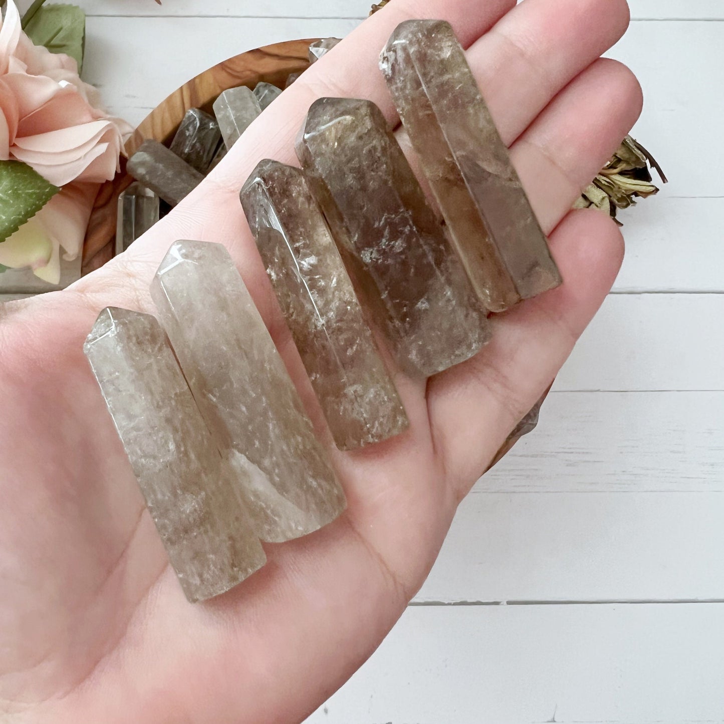 A set of six clear and 1-2" Smoky Quartz Point crystal points arranged on a white surface with pink roses in the background. The crystals have a polished, translucent appearance and range in height. Perfect for crystal grids, the roses add a soft, delicate contrast to these grounding stones.