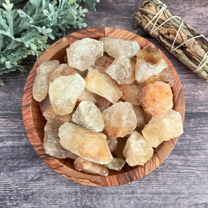 A wooden bowl filled with Raw Citrine Stones, known for activating the Solar Plexus Chakra, is placed on a wooden surface. Next to the bowl is a sage bundle, and some greenery can be seen in the upper left corner. The stones range in color from pale yellow to deeper amber tones.