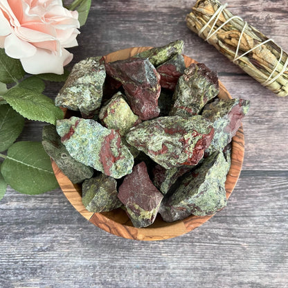 A wooden bowl filled with Raw Dragon Blood Jasper Stones sits on a rustic wooden surface. To the left, there are green leaves and a light pink rose. In the top right, a bundle of dried herbs is tied together with string, symbolizing inner strength and willpower.