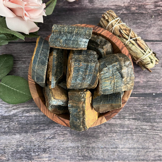 A wooden bowl filled with irregularly shaped, polished pieces of Raw Blue Tigers Eye Stones sits on a wooden surface. To the right of the bowl is a bundle of sage wrapped in cord, and to the left are green leaves and a light pink rose, enhancing the display's calming and healing effect.