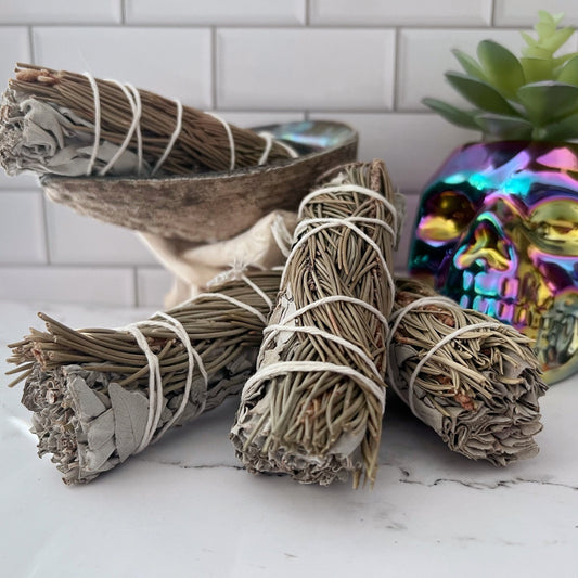 Bundles of Organic White Sage & Pine Smudge Sticks are tied with white string and placed on a white marble countertop. Behind them is a shell bowl and a colorful, iridescent skull decoration. A succulent plant in a white pot appears in the background against the white subway tile backsplash.