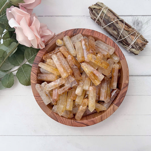 A wooden bowl filled with 1-2" Golden Healer Quartz Points in shades of yellow and clear sits on a white surface. To the left, there is a pink flower with leaves, and at the top right, an energy generator sage bundle wrapped with white string.