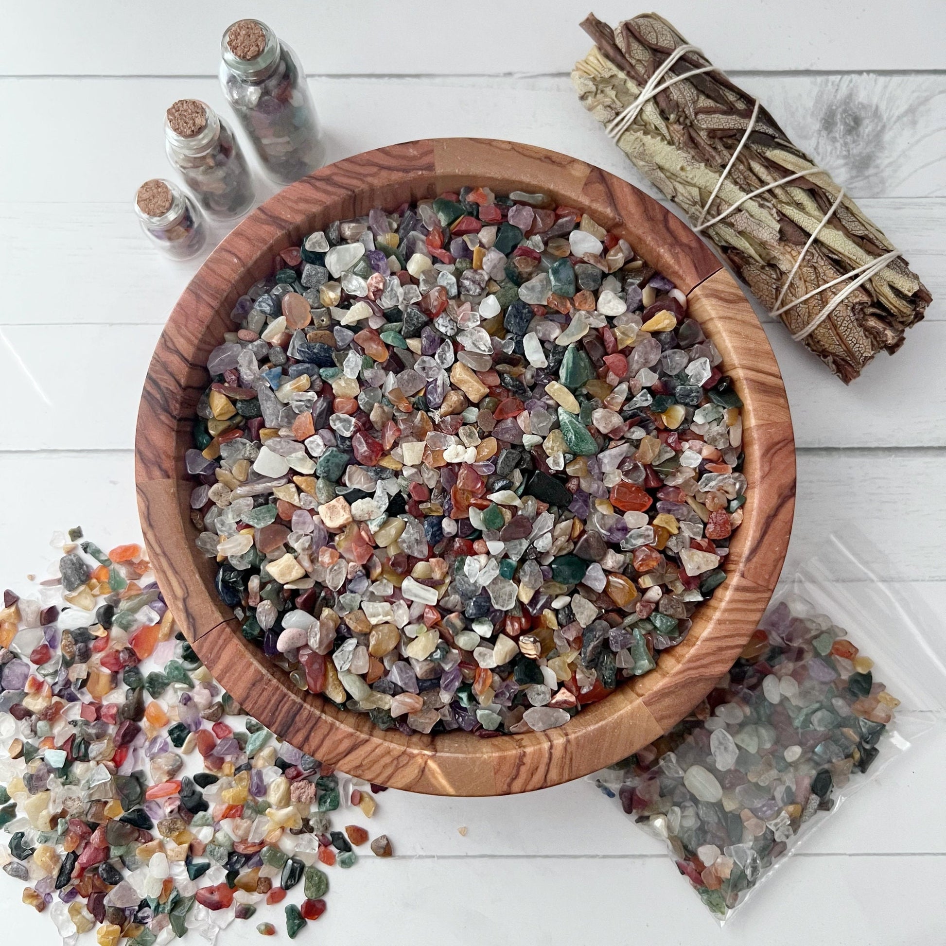 A bowl filled with various small, colorful polished stones sits on a white surface. Surrounding the bowl are Mixed Assorted Crystal Chips in small glass jars, two transparent bags containing more stones, and a bundle of sage tied with string. Perfect for metaphysical and crafting uses.