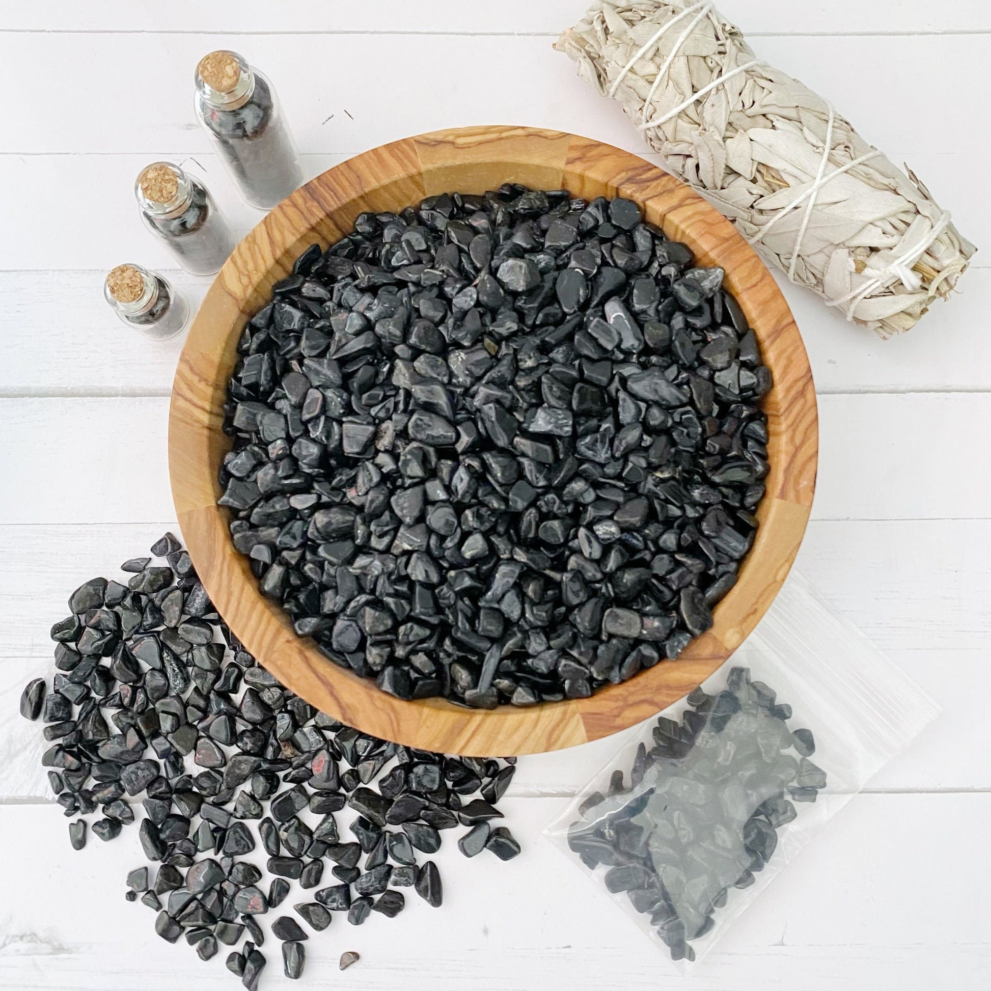 A wooden bowl filled with black stones, including Black Tourmaline Crystal Chips, is placed on a white surface. Surrounding the bowl are three small glass bottles with cork stoppers, a cluster of black stones scattered on the left, a bundle of white sage often used in spiritual practices, and a small plastic bag containing more protective crystals.