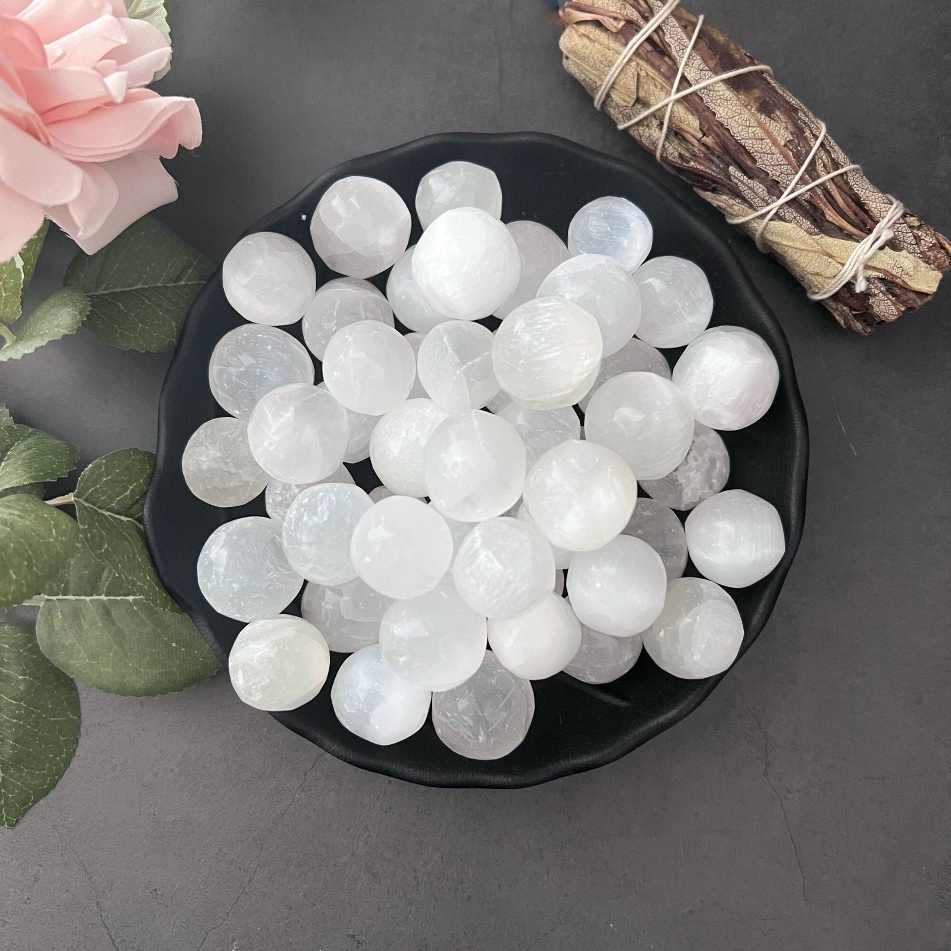 A black bowl filled with Mini Selenite Tumbled Spheres sits on a gray surface. To the left, there is a pink rose and some green leaves. In the upper right corner, there is a bundle of dried herbs wrapped with string, perfect for meditation and spiritual practice.