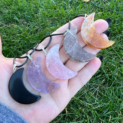 A picture features five crystal moons in different colors and stones: purple, gray, pink, yellow, and black. One black Gemstone Crescent Moon Crystal Pendant Charm is attached to a black cotton waxed cord necklace, while the rest are displayed on a white surface.