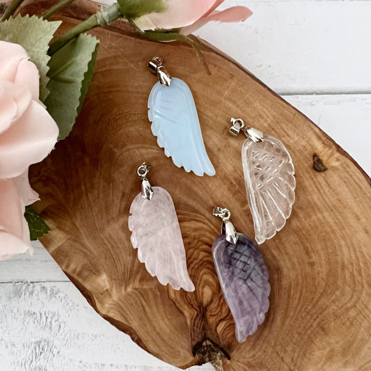 Four handcarved Angel Wing Crystal Pendants, made of light blue, clear, pink, and purple stones respectively, are displayed on a wooden surface. A pink flower with green leaves is positioned in the top left corner of the image. These Angel Wing Crystal Pendants are ideal emotional healing stones.