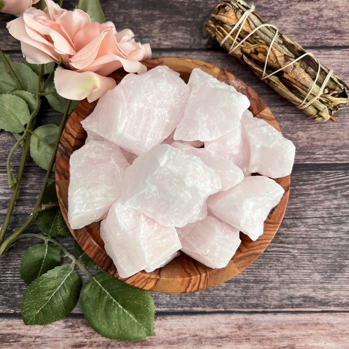 A wooden bowl filled with raw pink calcite stones sits on a wooden surface alongside pink calcite. Nearby, pale pink roses and a sage bundle wrapped with twine enhance the calming and serene atmosphere, perfect for cultivating inner peace and self-healing.