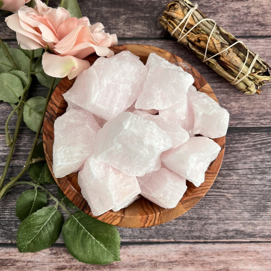 A wooden bowl filled with raw pink calcite stones sits on a wooden surface alongside pink calcite. Nearby, pale pink roses and a sage bundle wrapped with twine enhance the calming and serene atmosphere, perfect for cultivating inner peace and self-healing.