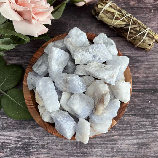 A wooden bowl filled with rough, white and light gray moonstones is placed on a wooden surface. To the left of the bowl, there's a pink rose and some green leaves. To the right, a sage bundle is visible along with a Raw Amblygonite Stone known for its soothing and healing properties due to its high lithium content.