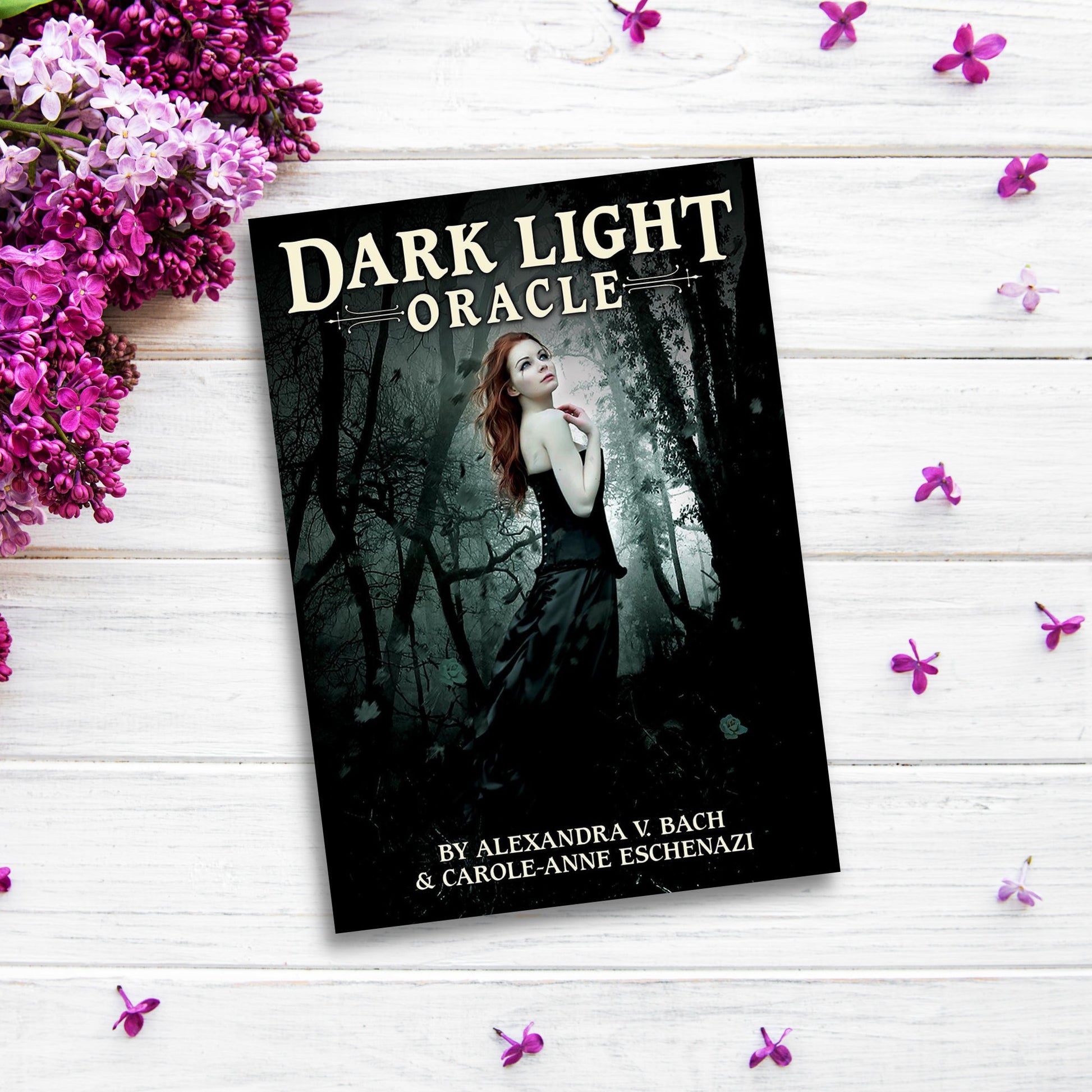 The product "Dark Light Oracle" is a book with a cover image of a woman in a dark forest, placed on a white wooden surface surrounded by lilac flowers and loose purple petals. Authored by Alexandra V. Bach and Carole-Anne Eschenazi, it promises profound healing and growth.