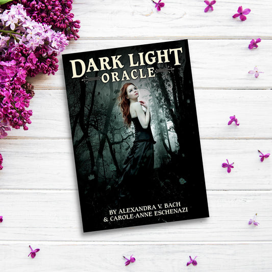 The product "Dark Light Oracle" is a book with a cover image of a woman in a dark forest, placed on a white wooden surface surrounded by lilac flowers and loose purple petals. Authored by Alexandra V. Bach and Carole-Anne Eschenazi, it promises profound healing and growth.