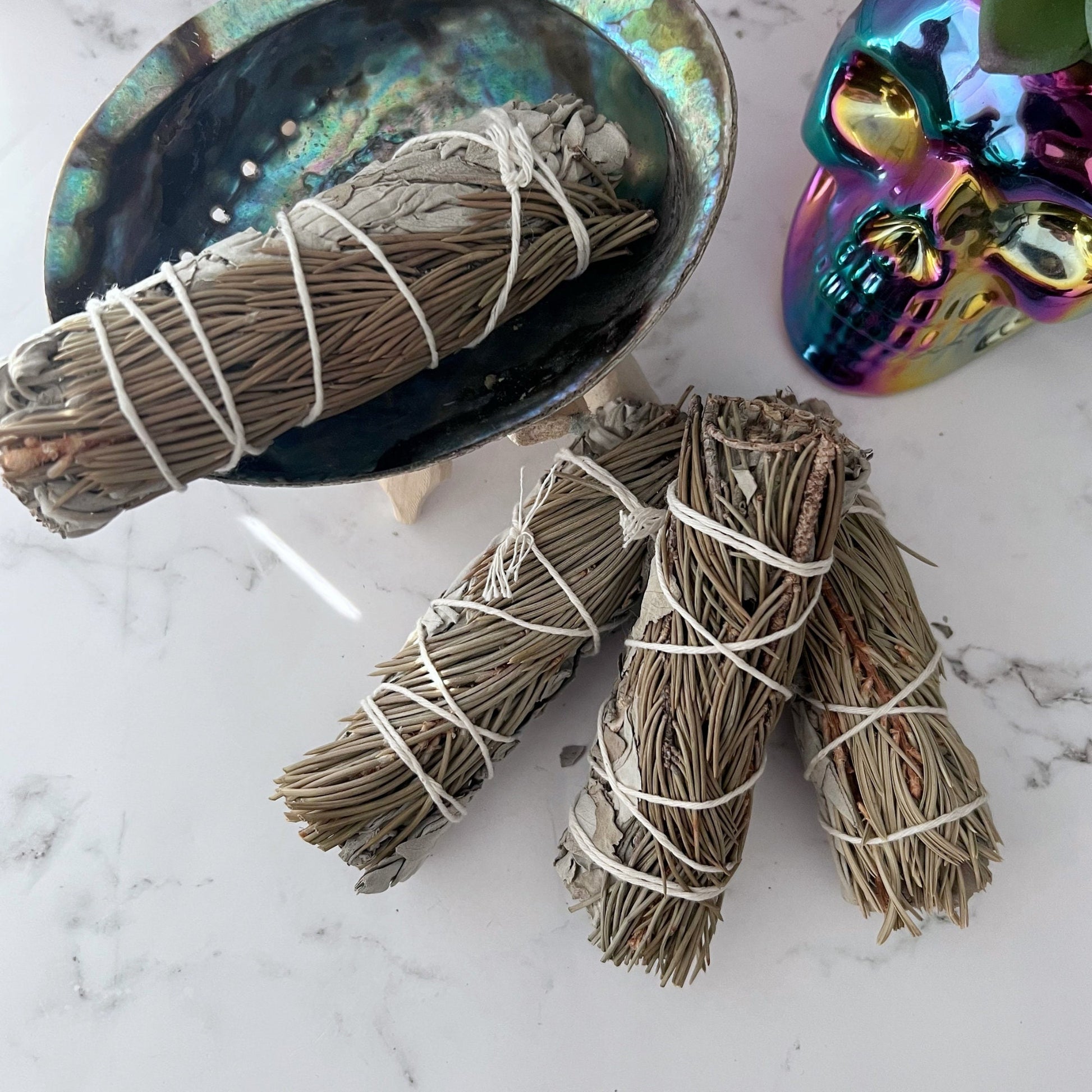 Bundles of Organic White Sage & Pine Smudge Sticks are tied with white string and placed on a white marble countertop. Behind them is a shell bowl and a colorful, iridescent skull decoration. A succulent plant in a white pot appears in the background against the white subway tile backsplash.