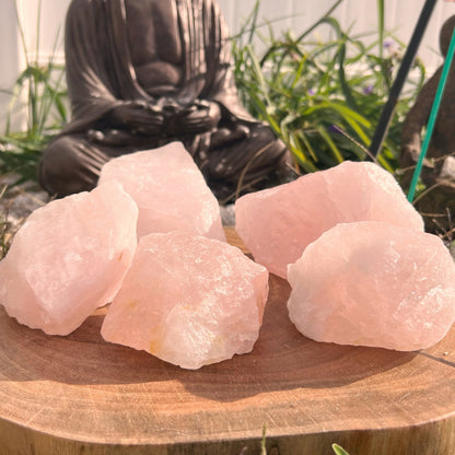 Five large raw rose quartz stones are displayed on a wooden slab outdoors. In the background, a bronze-colored statue of a meditative figure is surrounded by green plants, symbolizing emotional healing. The scene is bathed in warm sunlight, exuding an aura of unconditional love.