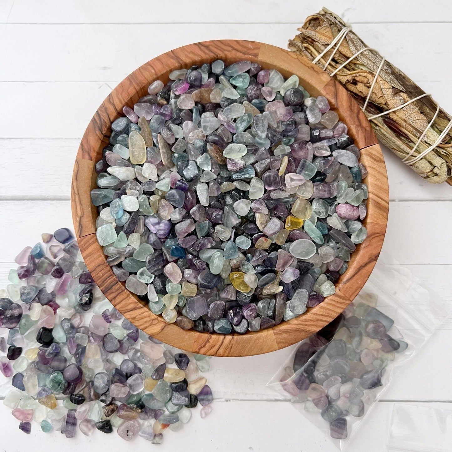 A wooden bowl filled with polished multicolored stones, including vibrant Rainbow Fluorite Crystal Chips, rests on a white surface. Some stones spill out of the bowl, alongside two small plastic bags containing more Rainbow Fluorite Crystal Chips. A bundle of dried sage for aura cleansing is placed beside the bowl.