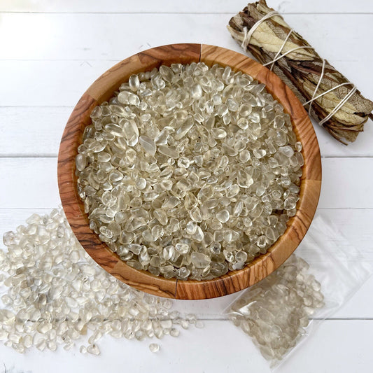 A wooden bowl filled with clear, polished stones sits on a white surface. Some tumbled and polished tiny stones are scattered beside the bowl. A small pack of Lemon Quartz Crystal Chips is nearby. In the top right corner, there is a bundle of dried leaves wrapped with string.