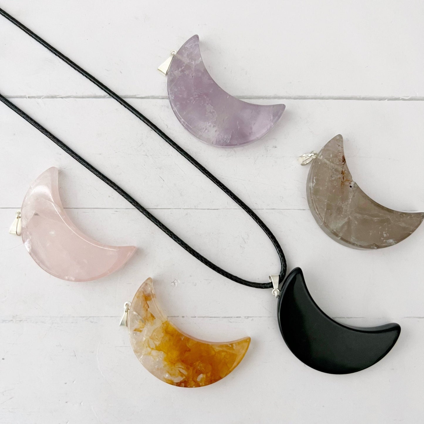 A picture features five crystal moons in different colors and stones: purple, gray, pink, yellow, and black. One black Gemstone Crescent Moon Crystal Pendant Charm is attached to a black cotton waxed cord necklace, while the rest are displayed on a white surface.