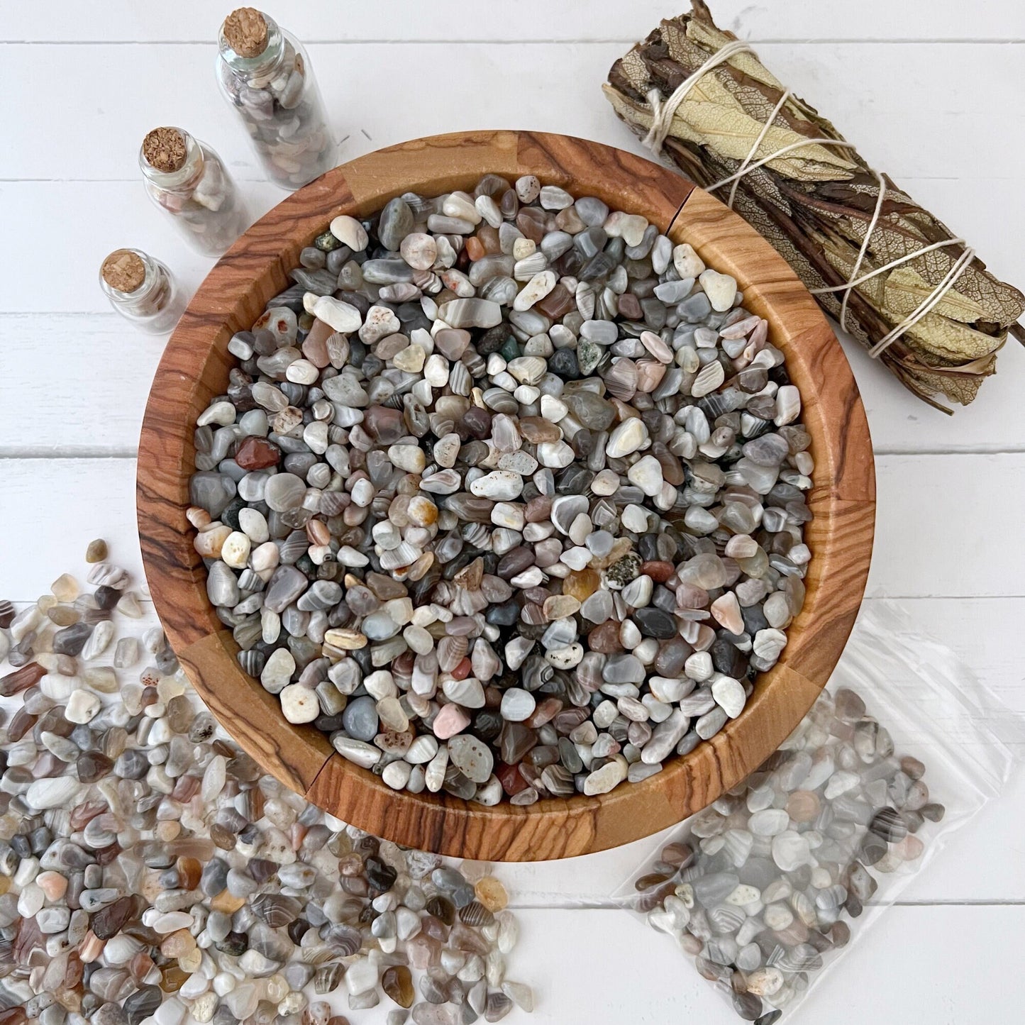 A wooden bowl filled with assorted small pebbles, including tumbled and polished stones, sits on a white surface. Surrounding it are three glass jars with cork stoppers containing more Botswana Agate Crystal Chips, a bundle of dried sage tied with string, and an open plastic bag spilling additional Botswana Agate Crystal Chips for metaphysical uses.