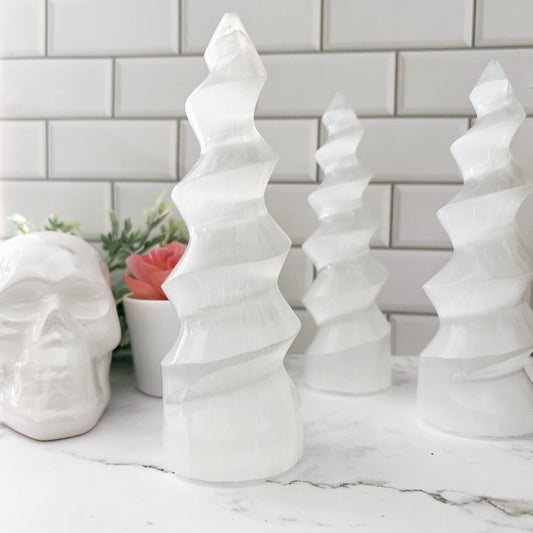 Four intricately designed white crystal trees are arranged on a marble surface. In the background, a 5-6" Selenite Crystal Twist Tower Skyscraper stands beside a white ceramic skull and a small succulent plant, all set against a backdrop of white subway tiles.