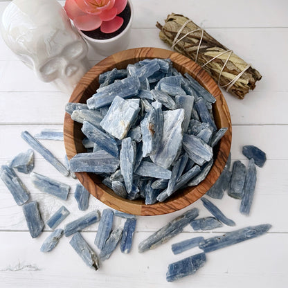 A wooden bowl filled with Raw Blue Kyanite Blades sits on a white surface, surrounded by scattered pieces of kyanite. Nearby, a small potted succulent, a bundle of sage, and a white skull-shaped decor piece are thoughtfully arranged to enhance psychic abilities and foster healing dreams.