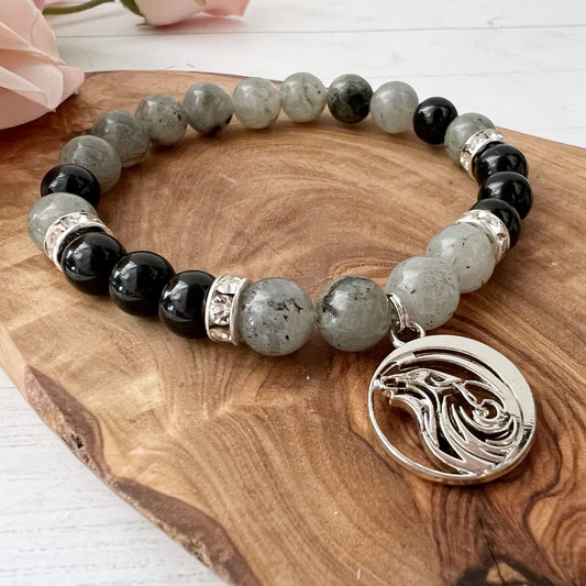 An 8mm Labradorite & Black Tourmaline Beaded Bracelet w/ Wolf Charm is displayed on a wooden surface. The bracelet features silver spacers and a silver pendant with an intricate design. Pink rose petals are partially visible in the background.