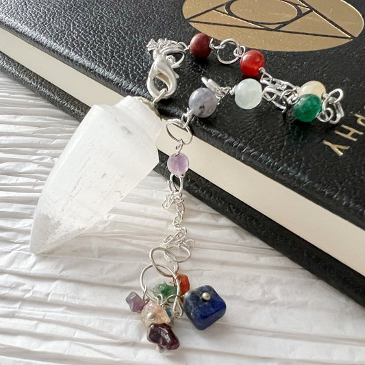 A Selenite Teardrop Chakra Pendulum with a clear point and a chain adorned with various colorful stones is resting on a closed black book with a gold-embossed symbol on the cover. The background features a white, textured surface, adding an elegance that enhances any spiritual practice.