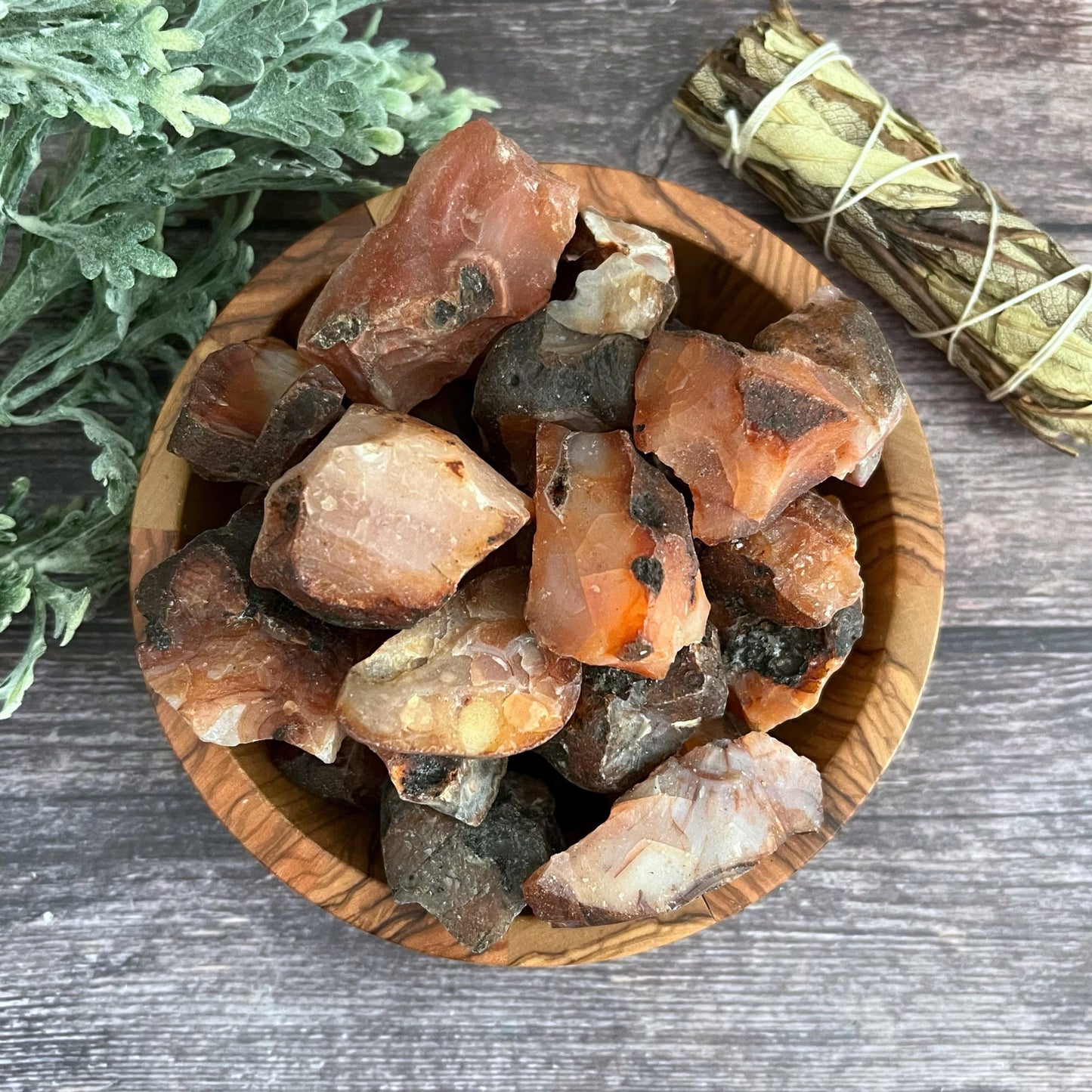 A wooden bowl full of rough, unpolished crystal stones sits on a wooden surface. To the left of the bowl is a sprig of leafy green plant, and above the bowl is a bundle of sage wrapped in twine. Among the crystals, vibrant Raw Carnelian Stones stand out, radiating motivation and endurance.