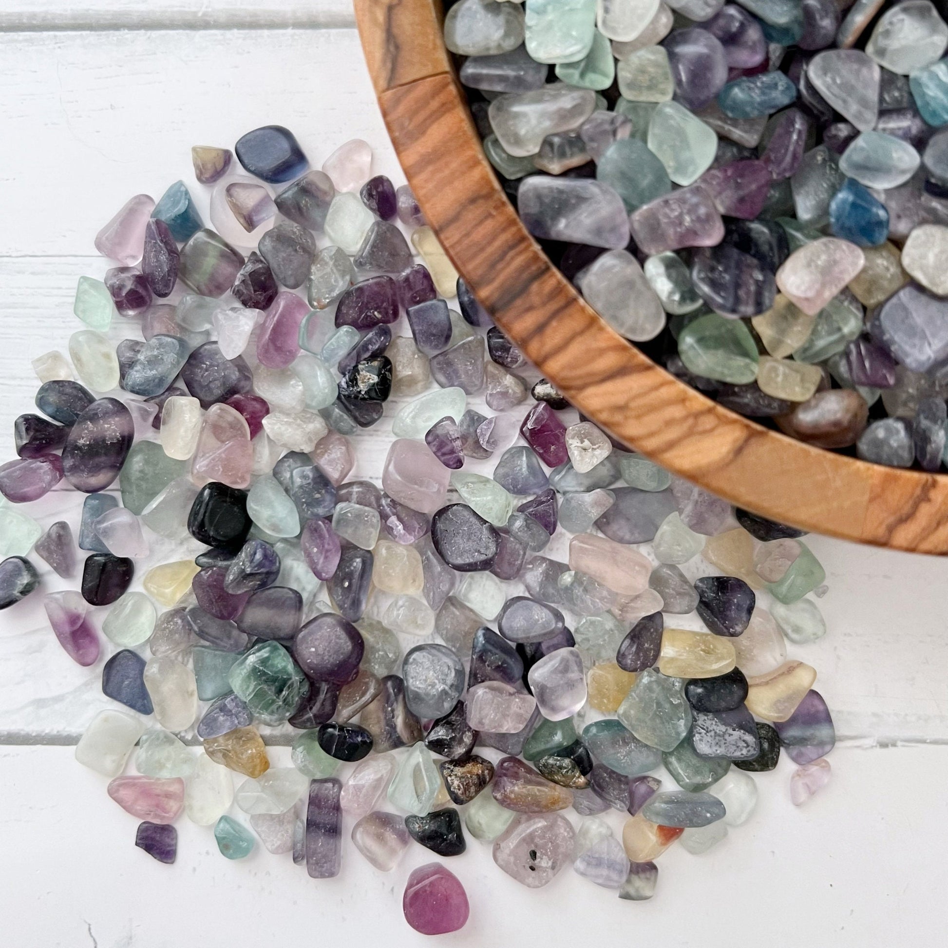 A wooden bowl filled with polished multicolored stones, including vibrant Rainbow Fluorite Crystal Chips, rests on a white surface. Some stones spill out of the bowl, alongside two small plastic bags containing more Rainbow Fluorite Crystal Chips. A bundle of dried sage for aura cleansing is placed beside the bowl.