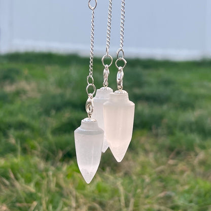 A Selenite Teardrop Chakra Pendulum with a clear point and a chain adorned with various colorful stones is resting on a closed black book with a gold-embossed symbol on the cover. The background features a white, textured surface, adding an elegance that enhances any spiritual practice.